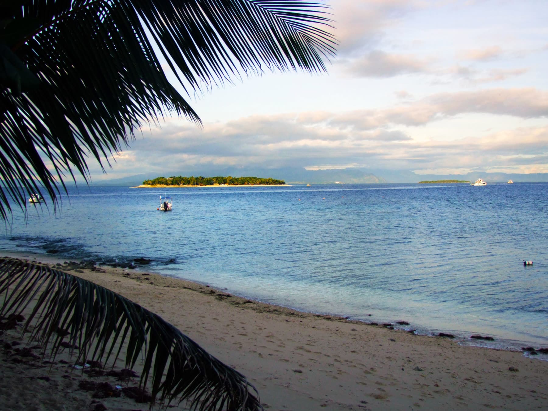 A sandy beach and palm fronds looking across the water toward the Mamanuca Islands in Fiji